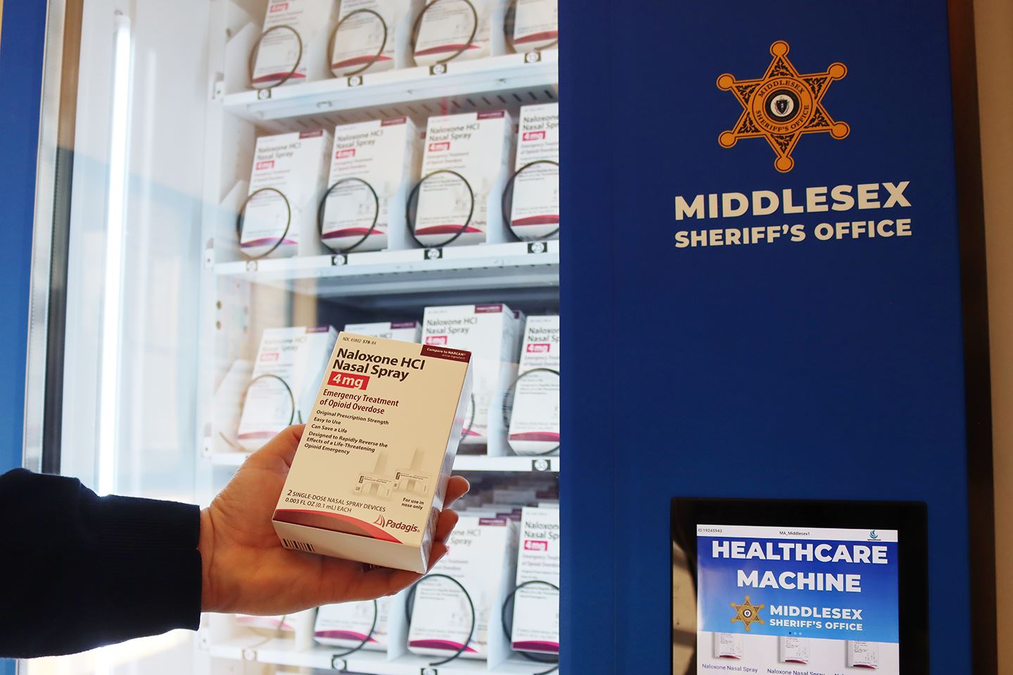 A naloxone kit is held in front of a naloxone vending machine.