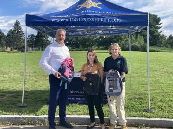 Sheriff Koutoujian, Family Resource and Outreach Coordinator Lili Bonilla and Education Director Lisa Aubin stand outside, in front of a tent holding backpacks.