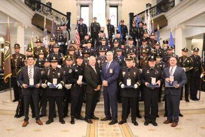 Award recipients and officials stand in front of a large group of honor guard members at the base of the Grand Staircase inside the Massachusetts State House.